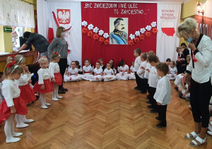 Dziewczynki w stojach biało-czerwonych oraz chłopcy ubrani na galowo stoją naprzeciwko siebie i śpiewają piosenkę o ułanach.